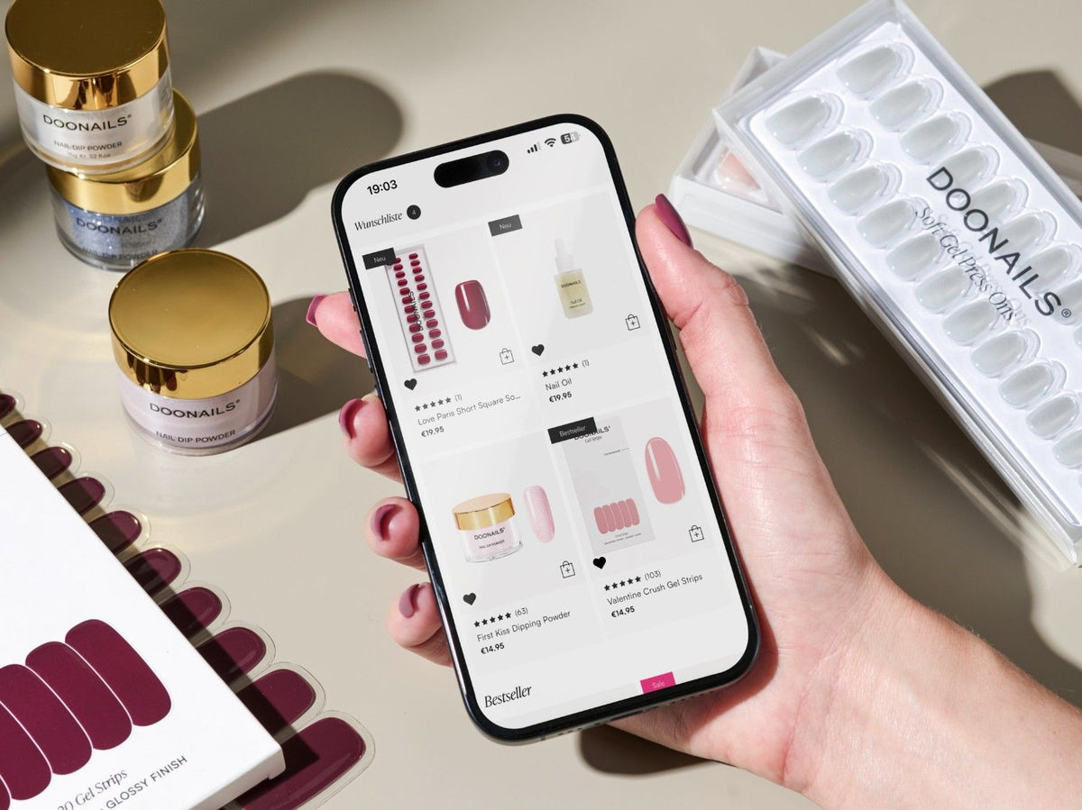 Ein Handy auf dem die Doonails App gezeigt wird, wird in der Hand gehalten. Im Hintergrund sind viele Doonails Produkte.