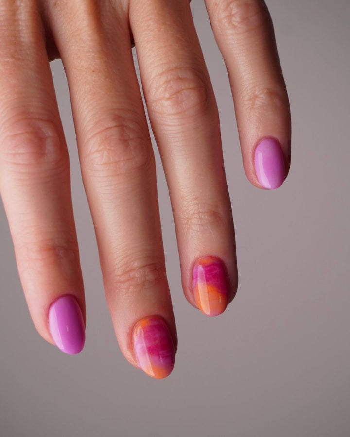 Hand mit Dipping Powder Nageldesign in Pink und Orange auf einem hellgrauen Hintergrund. 