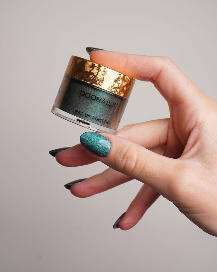 Hand hält eine glitzernde, grün-blaue Dipping Powder Dose in der Hand. 