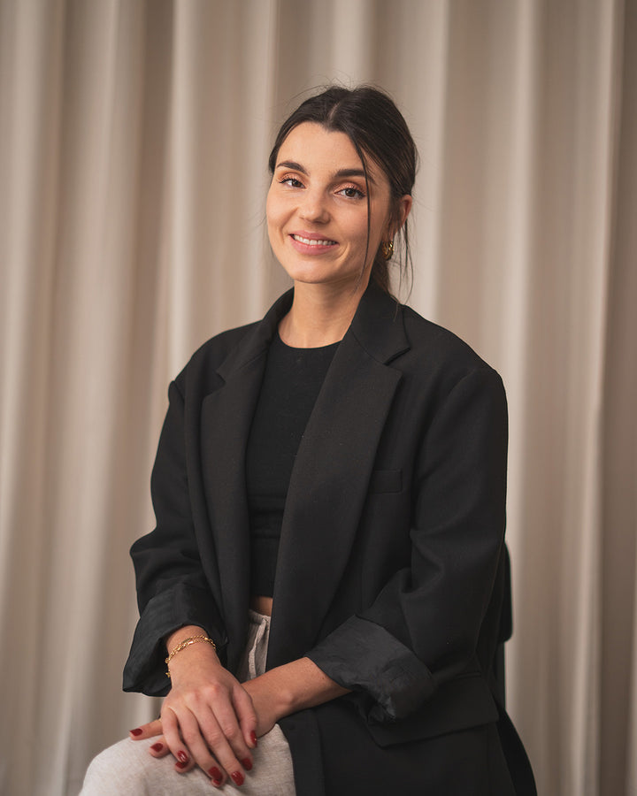 Gründerin Elisa mit schwarzem Blazer und roten Nägeln. Hintergrund ist ein beigefarbener Vorhang.