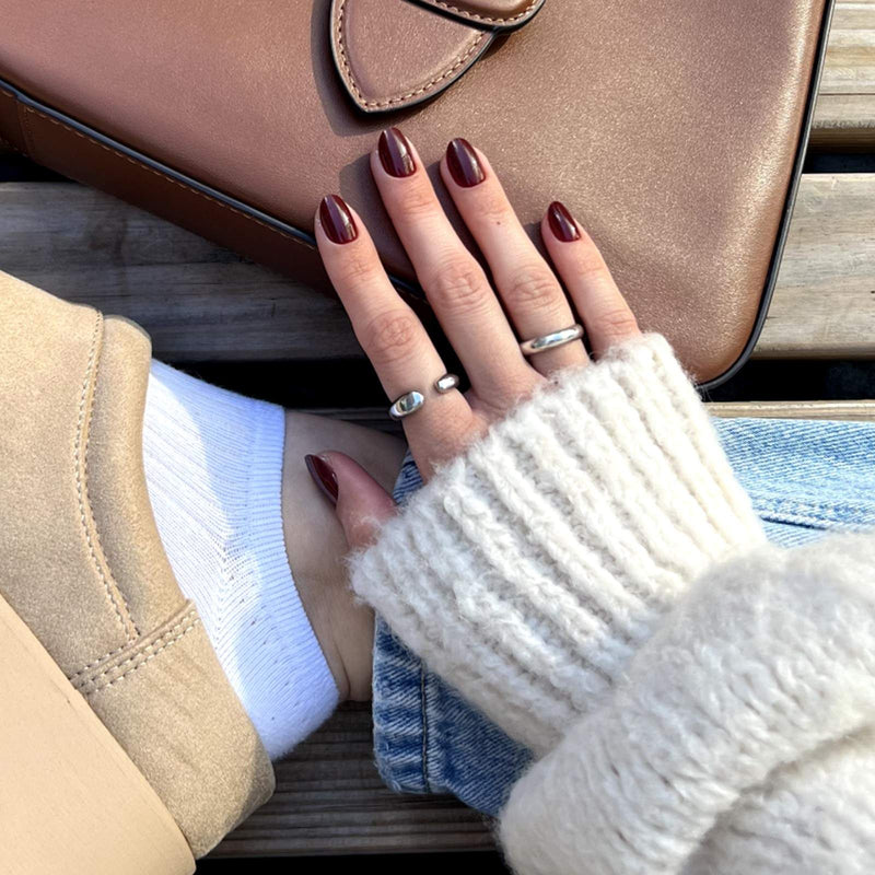 Eine Hand mit dunkelroten Nägeln liegt auf einer braunen Tasche. Getragen wird ein beiger Pullover und beigefarbene Schuhe.