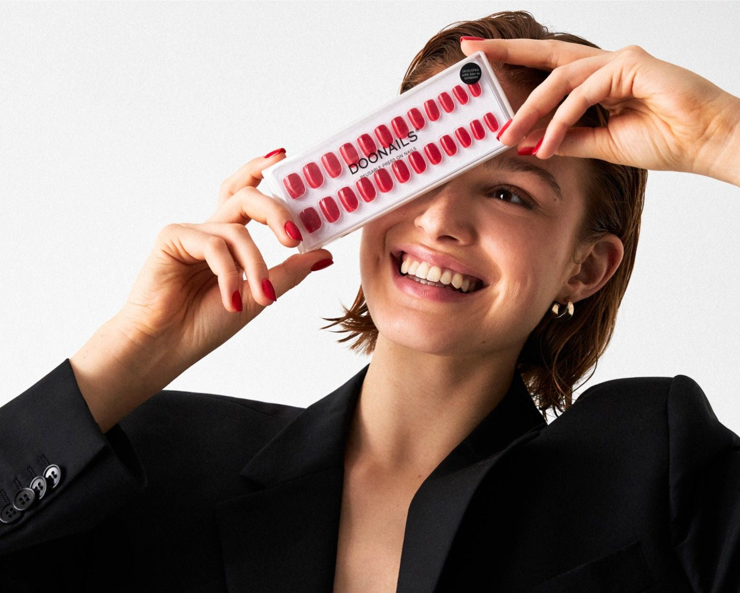 Lächelnde Frau hält eine Packung roter Sotf Gel Press Ons der Marke Doonails vor ihr Gesicht, trägt schwarzen Blazer und rote Press Ons.