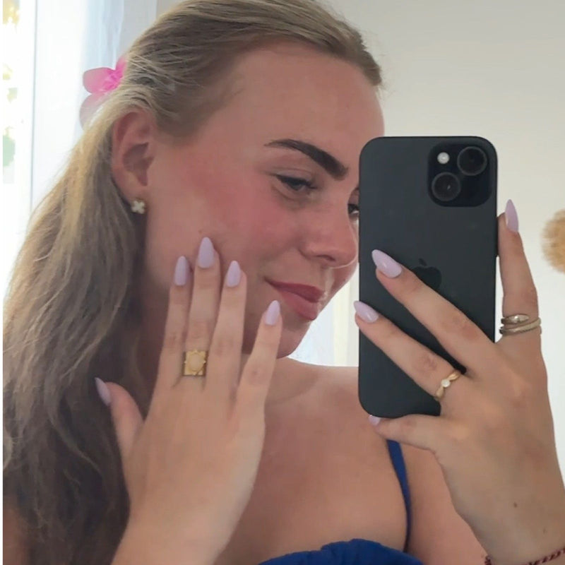 Blondes Mädchen macht Spiegel-Selfie, hält ihre Hand an die Backe und zeigt ihre pastell-lila Soft Gel Press Ons.