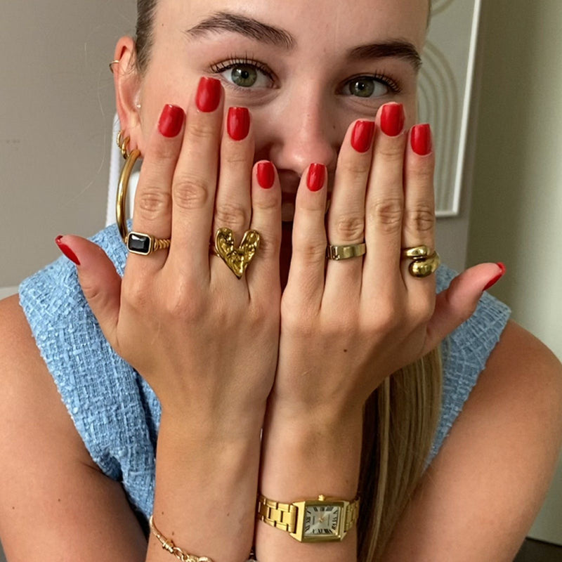 Lächelndes Mädchen mit grünen Augen hält sich ihre Hände mit roten Dipping-Powder-Nägeln und goldenen Ringen vor ihr Gesicht.